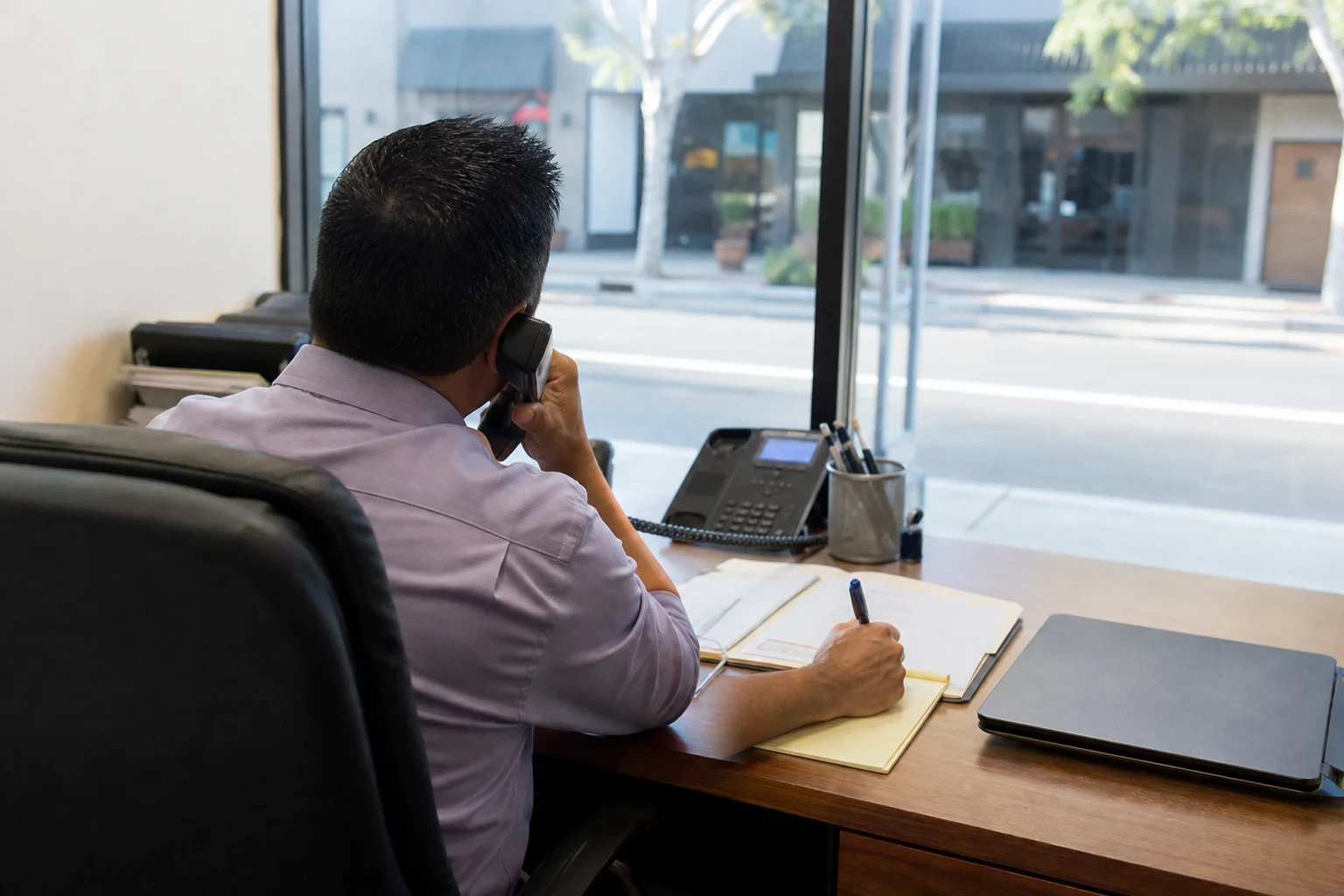 Agent in a lavender shirt on a desk phone, writing on a yellow notepad beside an open file folder and closed laptop, facing a window onto a quiet street
